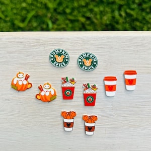 May include: A collection of fall-themed decorative accessories, including pumpkin spice logo pins, pumpkin-shaped characters, and coffee cup designs. The color palette features orange, red, white, and green, perfect for autumn-themed crafts and decorations.