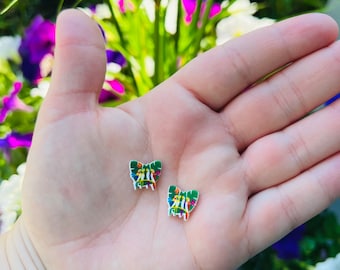 May include: Two small butterfly-shaped earrings with a colorful floral design. The earrings have a white outline and are held in a hand against a background of green foliage and flowers.