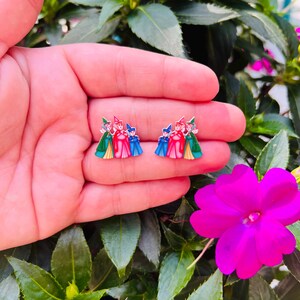 May include: A pair of enamel earrings featuring a design of three figures in colorful attire. The earrings are held in a hand, set against a backdrop of green foliage and vibrant pink flowers.