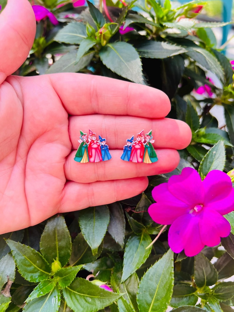 May include: A pair of enamel earrings featuring a design of three figures in colorful attire. The earrings are held in a hand, set against a backdrop of green foliage and vibrant pink flowers.