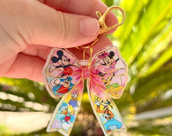 May include: A pink bow-shaped keychain featuring classic Disney characters like Mickey and Minnie Mouse, Donald Duck, and Goofy. The keychain has a gold-colored clasp and ring, and the bow has a gold outline.