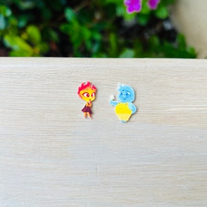 May include: Two cartoon character figures. One figure is red and orange with a dark purple dress. The other figure is blue and yellow, holding a white object. The figures are on a light wood-grain surface.