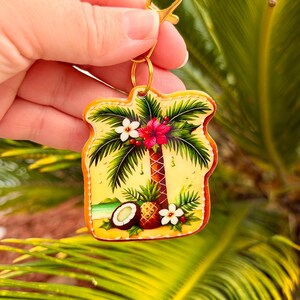 May include: A tropical-themed keychain featuring a palm tree, flowers, and a coconut on a yellow background. The keychain has a gold-colored clasp and ring. The design includes a beach scene with a pineapple and a red hibiscus flower.