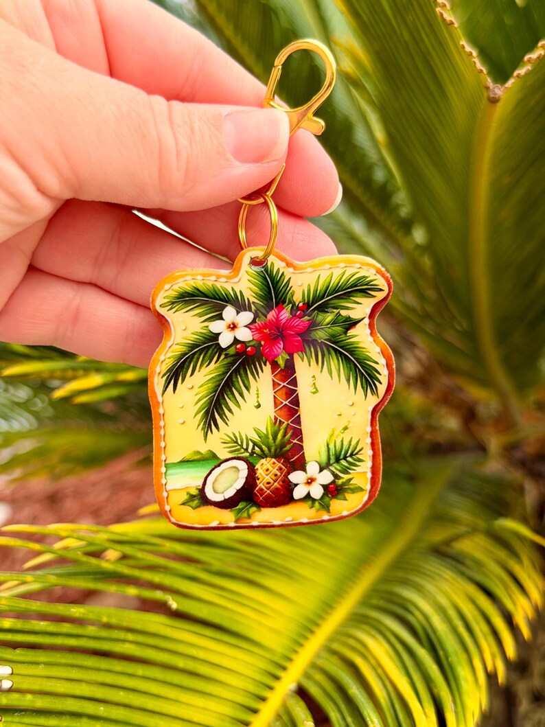 May include: A tropical-themed keychain featuring a palm tree, flowers, and a coconut on a yellow background. The keychain has a gold-colored clasp and ring. The design includes a beach scene with a pineapple and a red hibiscus flower.