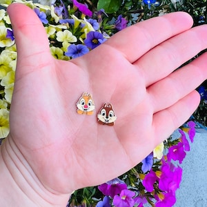 May include: Two small, cartoon chipmunk pins. The chipmunks are brown and white with black eyes and noses. The pins are on a hand with a light pink background.