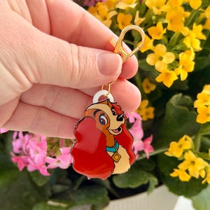 May include: A colorful keychain featuring a cartoon dog with red hair, a blue collar, and a gold clasp. The dog is smiling. The keychain is held in front of a background of pink and yellow flowers.