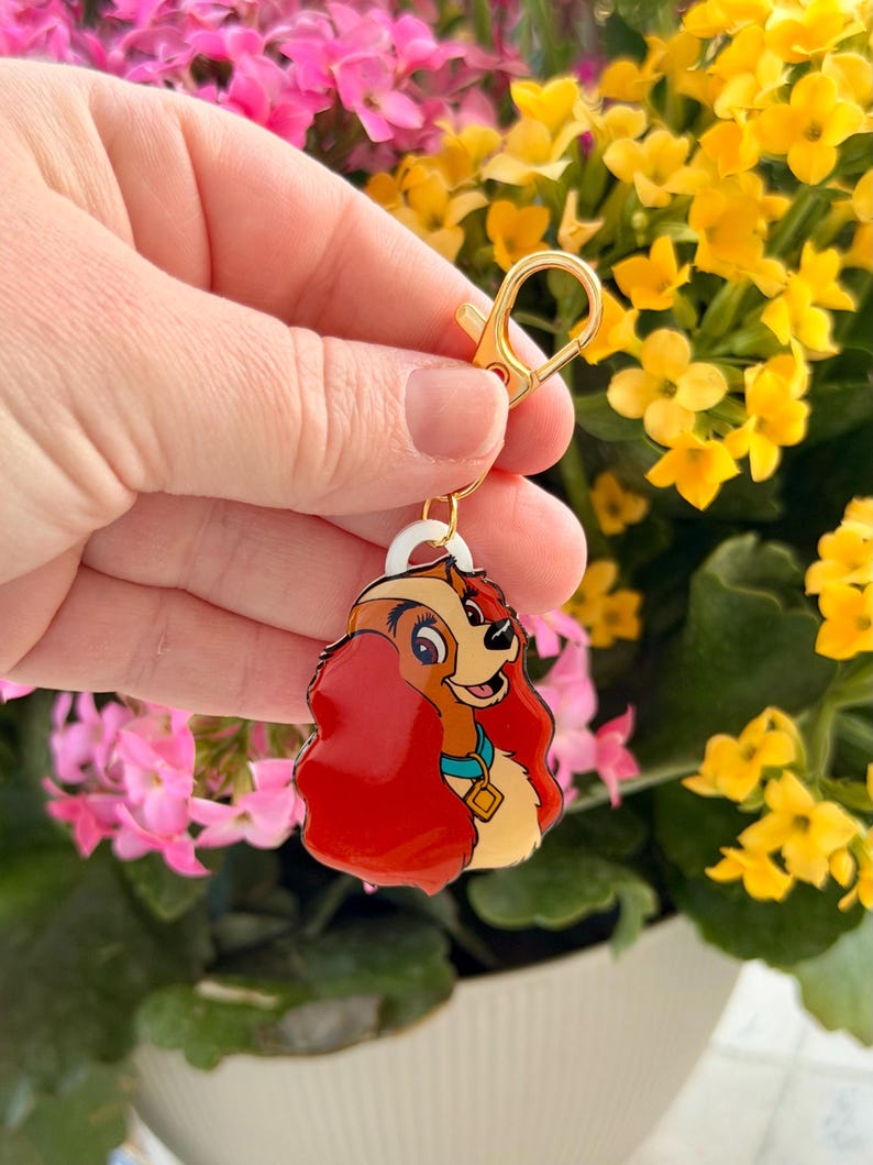May include: A colorful keychain featuring a cartoon dog with red hair, a blue collar, and a gold clasp. The dog is smiling. The keychain is held in front of a background of pink and yellow flowers.
