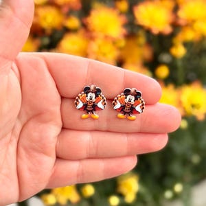 May include: A pair of festive stud earrings featuring a cartoon character dressed as a turkey. The earrings have a black, brown, orange, and white color scheme. The earrings are held in a hand against a blurred background of yellow flowers.