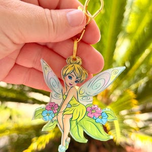 May include: A colorful keychain featuring a cartoon fairy with yellow hair, a green dress, and translucent wings. The fairy is seated on a green leaf with pink and blue flowers. The keychain has a gold-colored clasp.