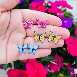 May include: Three pairs of butterfly earrings. One pair is pink with a swirl design, one pair is blue, and one pair is yellow with a floral design.