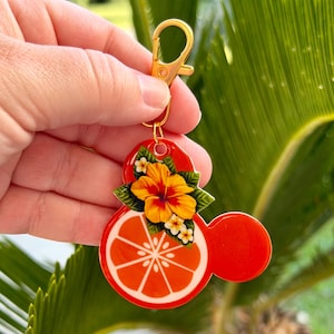 Może przedstawiać: Czerwony i pomarańczowy brelok do kluczy ze złotym zapięciem. Brelok przedstawia plaster pomarańczy z białym środkiem i ż&oacute;łto-pomarańczowym kwiatem. Projekt ma kształt głowy myszy.