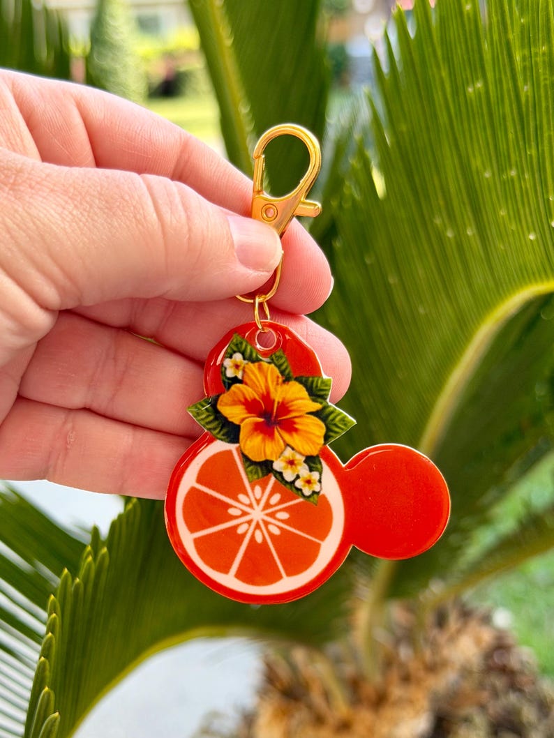 Może przedstawiać: Czerwony i pomarańczowy brelok do kluczy ze złotym zapięciem. Brelok przedstawia plaster pomarańczy z białym środkiem i ż&oacute;łto-pomarańczowym kwiatem. Projekt ma kształt głowy myszy.
