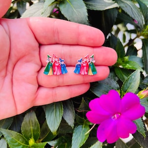 May include: A pair of colorful stud earrings featuring a cartoon design of three figures in red, blue, and green outfits. The earrings are held in a hand, with a background of green leaves and a bright pink flower.