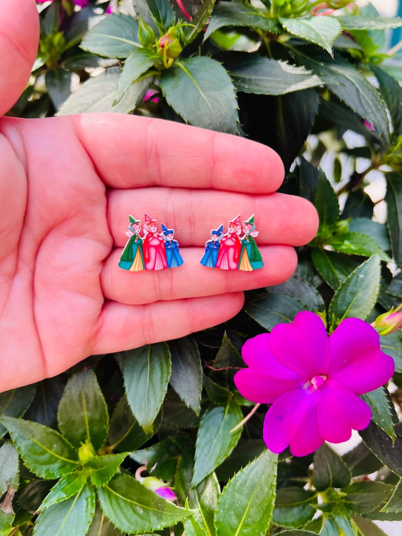 May include: A pair of colorful stud earrings featuring a cartoon design of three figures in red, blue, and green outfits. The earrings are held in a hand, with a background of green leaves and a bright pink flower.