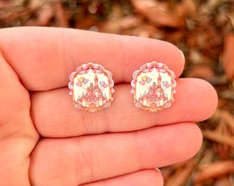 May include: A pair of stud earrings featuring a castle design. The earrings have a pink and white color scheme with a decorative border. The castle design includes a pink roof and a white facade, with small butterflies.