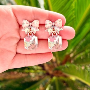 May include: A pair of dangle earrings featuring a bow with a floral pattern in pink and white. Below the bow is a rectangular charm with a pink castle design, pink clouds, and a Mickey Mouse head. The earrings are held in a hand.