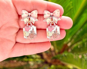May include: A pair of dangle earrings featuring a bow with a floral pattern in pink and white. Below the bow is a rectangular charm with a pink castle design, pink clouds, and a Mickey Mouse head. The earrings are held in a hand.