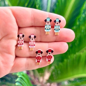 May include: Six pairs of colorful enamel stud earrings. Each earring features a cartoon character dressed as a toy soldier. The earrings are in shades of red, pink, blue, and gold. The earrings are small and detailed, with a glossy finish.