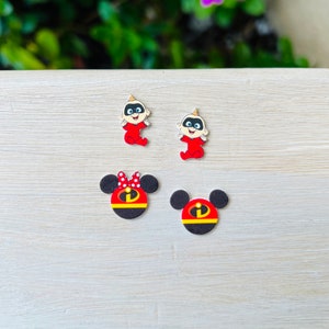 May include: Four small, colorful, cartoon character earrings. Two earrings feature a red and black superhero character with a red bow. The other two earrings feature a baby in a red and black suit.