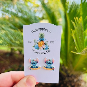 May include: A pair of Stitch stud earrings on a white card with the text "Pineapples & Pixie Dust Co.". The earrings feature a blue alien character with floral accents. The card has a pineapple illustration.