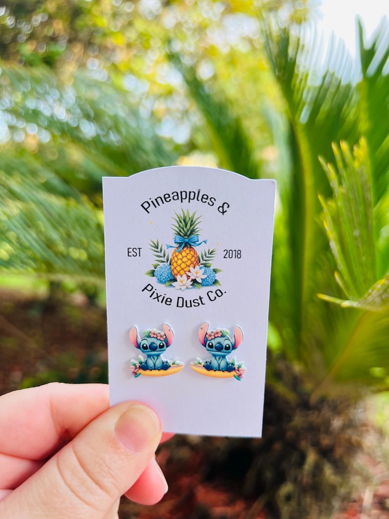 Op de afbeelding: Stitch-oorbellen op een witte kaart. De oorbellen tonen een blauw alien-personage met een bloemenkroon. De kaart heeft de tekst "Pineapples & Pixie Dust Co." en een ananasafbeelding.