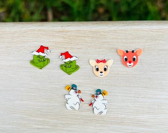 May include: A set of six flat decorative charms featuring holiday characters: two green figures wearing red Santa hats, one tan deer with a red bow, one orange deer with a red nose, and two white snowmen wearing blue hats and scarves.