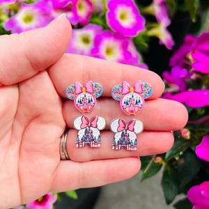 May include: A pair of pink and white Minnie Mouse ears with a pink castle design. The ears are decorated with pink bows and flowers.