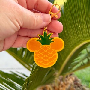 May include: A yellow and green pineapple-shaped keychain with a gold-colored clasp. The pineapple design incorporates Mickey Mouse ears. The keychain is held up against a background of green palm leaves.