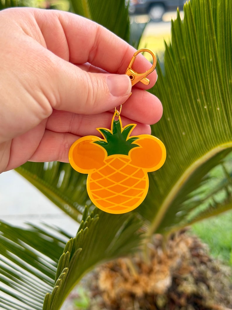 May include: A yellow and green pineapple-shaped keychain with a gold-colored clasp. The pineapple design incorporates Mickey Mouse ears. The keychain is held up against a background of green palm leaves.