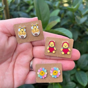 May include: A collection of colorful stud earrings featuring cartoon characters and floral designs. The earrings are displayed on small, brown cardboard cards. The designs include a bear in a honey pot, a bear in a red outfit, and colorful flower designs.