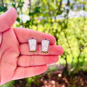 May include: A pair of novelty earrings shaped like cartoon doors. Each earring is white with a pink top, a red accent, and a yellow and black base. The earrings are held in a hand against a blurred green background.