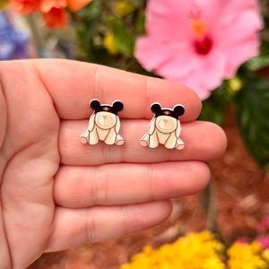 May include: A pair of small, cream-colored bear-shaped earrings with black hats and gold accents. The earrings are held in a hand, with a blurred background of flowers in shades of pink, orange, and yellow.