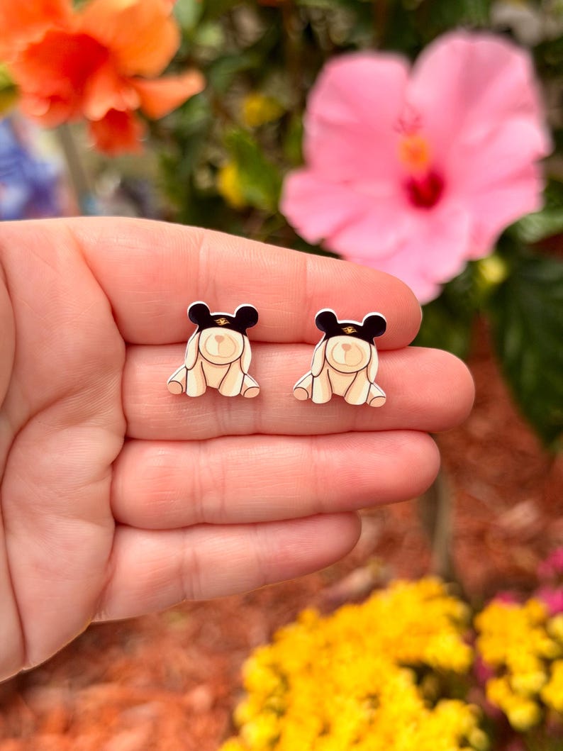 May include: A pair of small, cream-colored bear-shaped earrings with black hats and gold accents. The earrings are held in a hand, with a blurred background of flowers in shades of pink, orange, and yellow.