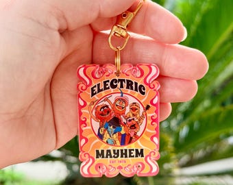 May include: A rectangular keychain with a gold-colored clasp. The keychain features the words "ELECTRIC MAYHEM" in orange and red, with a graphic of the Muppets band. The text "EST. 1975" is also visible. The background is a vibrant orange.