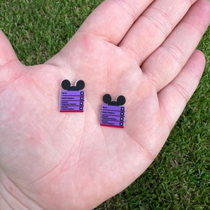 May include: Two purple and red Mickey Mouse-shaped pins with a list of Disney parks and their locations. The pins are on a hand.