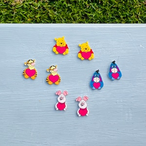 May include: A set of eight colorful cartoon character earrings featuring Winnie the Pooh, Tigger, Piglet, and Eeyore. Each character is holding a red heart.
