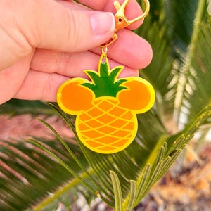 May include: A yellow and green pineapple-shaped keychain with a gold-colored clasp. The pineapple design incorporates Mickey Mouse ears. The keychain is held against a backdrop of green foliage.