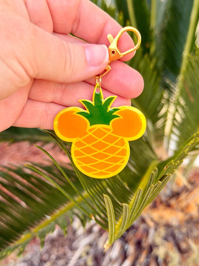 May include: A yellow and green pineapple-shaped keychain with a gold-colored clasp. The pineapple design incorporates Mickey Mouse ears. The keychain is held against a backdrop of green foliage.