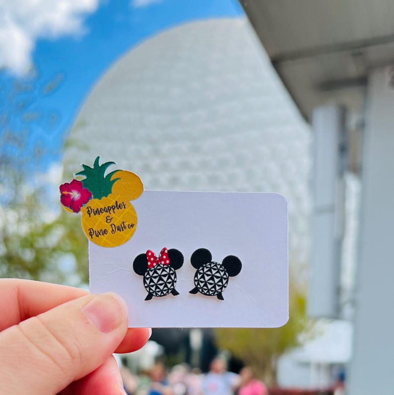 May include: A pair of black and white Mickey Mouse-shaped earrings with geometric patterns. One earring has a red bow. The earrings are displayed on a white card with a pineapple and flower sticker that reads "Pineapples & Pixie Dust Co."