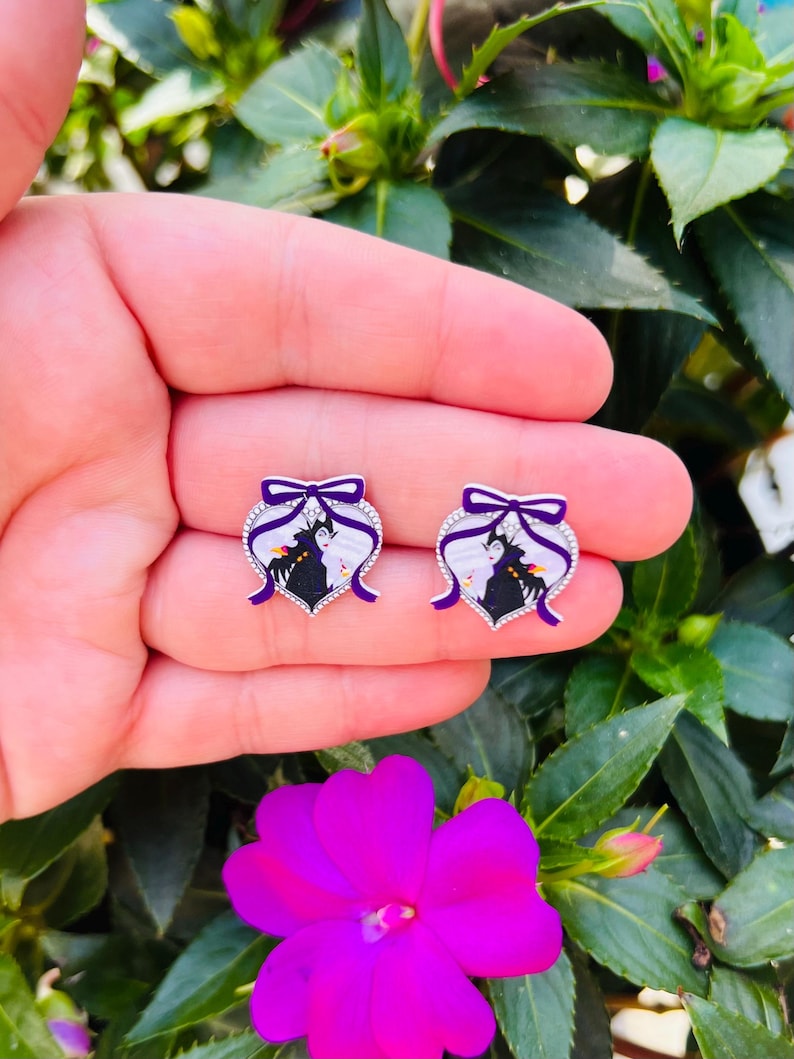 Puede incluir: Un par de pendientes de plata en forma de coraz&oacute;n con un lazo morado y una imagen en blanco y negro de Mal&eacute;fica y Aurora de la pel&iacute;cula de Disney La Bella Durmiente.