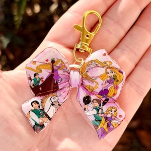 May include: A pink bow-shaped keychain with a gold-colored clasp. The bow features illustrations of characters from a fairytale, including a princess with long blonde hair. The bow has a glittery, textured finish.