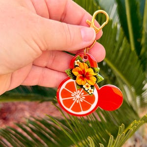 Może przedstawiać: Czerwony i pomarańczowy brelok do kluczy ze złotym zapięciem. Brelok ma wz&oacute;r plastra owocu cytrusowego z akcentem kwiatowym i liściastym. Projekt ma kształt znanej postaci z kresk&oacute;wek.