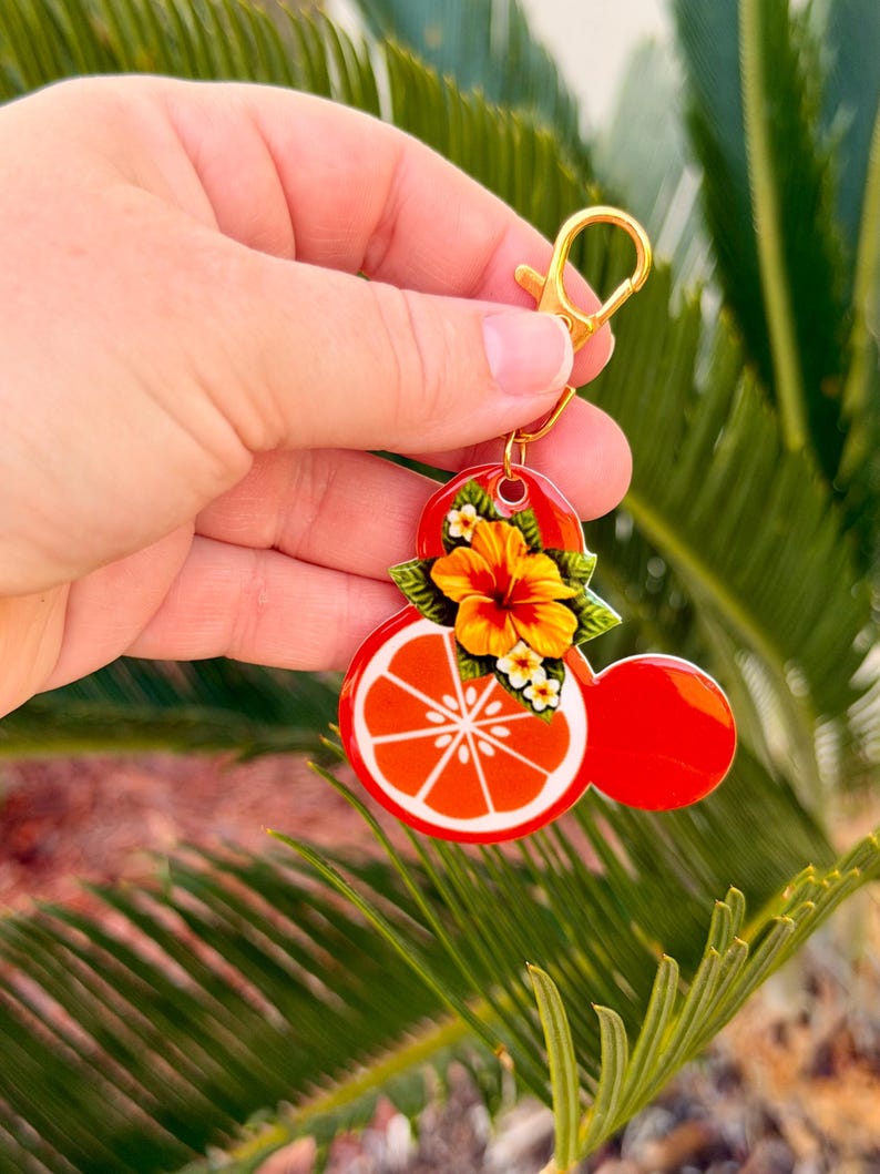 Może przedstawiać: Czerwony i pomarańczowy brelok do kluczy ze złotym zapięciem. Brelok ma wz&oacute;r plastra owocu cytrusowego z akcentem kwiatowym i liściastym. Projekt ma kształt znanej postaci z kresk&oacute;wek.