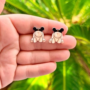 May include: A pair of bear-shaped stud earrings. Each earring features a cream-colored bear with a black hat and gold band. The earrings are displayed against a blurred green background.