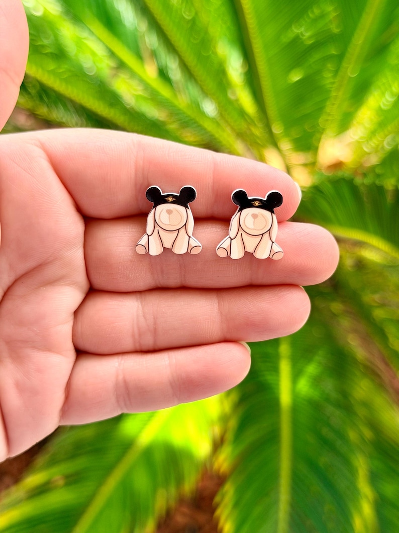 May include: A pair of bear-shaped stud earrings. Each earring features a cream-colored bear with a black hat and gold band. The earrings are displayed against a blurred green background.