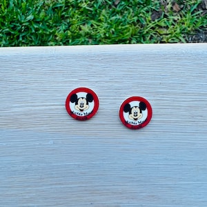 Op de afbeelding: Twee ronde Mickey Mouse-knopen. Elke knoop heeft Mickey's gezicht in het midden, omgeven door een rode rand. De tekst "Mickey Mouse Club" staat onder Mickey's gezicht. De knopen liggen op een lichtgekleurd houten oppervlak.