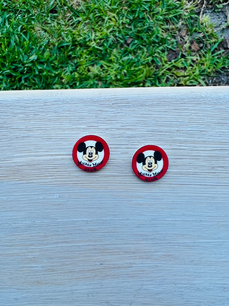 Op de afbeelding: Twee ronde Mickey Mouse-knopen. Elke knoop heeft Mickey's gezicht in het midden, omgeven door een rode rand. De tekst "Mickey Mouse Club" staat onder Mickey's gezicht. De knopen liggen op een lichtgekleurd houten oppervlak.