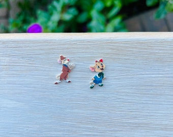 May include: Two colorful enamel pins featuring cartoon mice characters. One mouse wears a brown suit and hat, holding a magnifying glass. The other mouse is in a blue outfit with a red hat. The pins are set against a light wood grain surface.