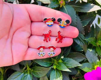 May include: A collection of colorful stud earrings featuring cartoon character designs. The earrings include Mickey Mouse, a red horse, and a cowgirl. The earrings are displayed against a background of green foliage and pink flowers.