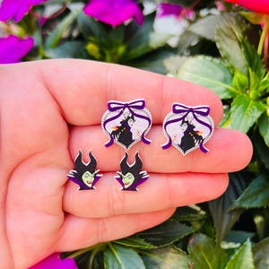 Puede incluir: Un conjunto de cuatro pendientes con la Reina Malvada de Blancanieves. Dos pendientes son en forma de coraz&oacute;n con un lazo morado y un fondo blanco, y los otros dos pendientes muestran la cara de la Reina Malvada.